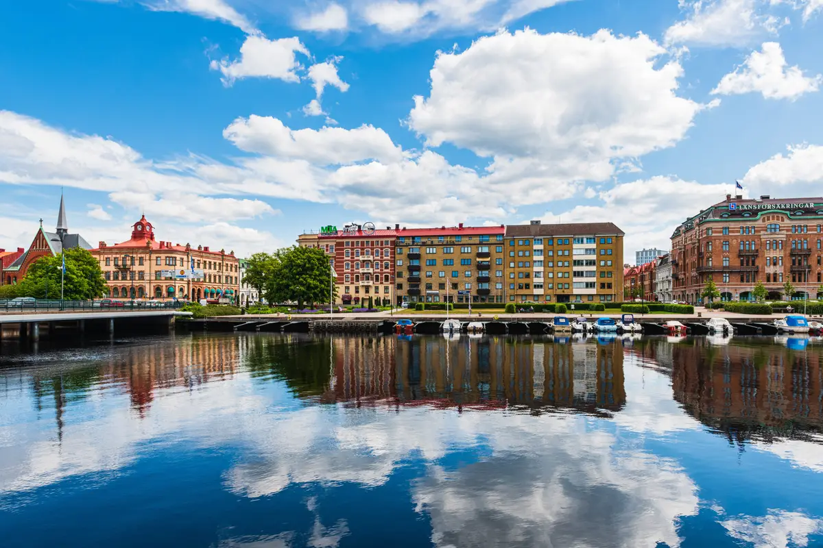 Colorful buildings lining the tranquil riverfront in Halmstad, Sweden, reflecting beautifully on the water surface, with boats moored along the jetty.