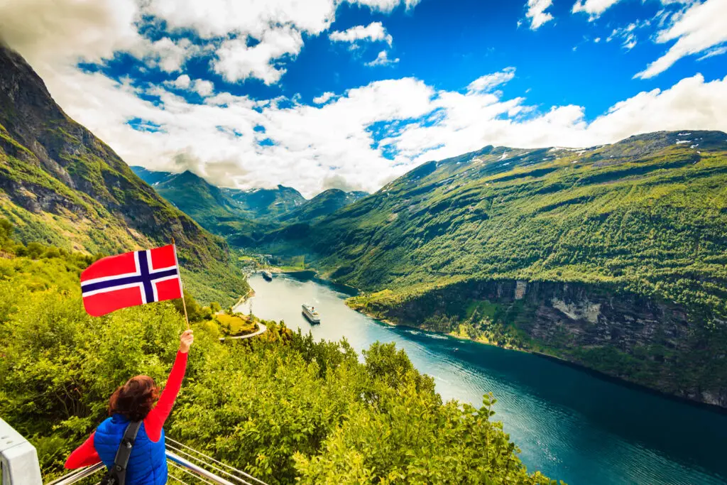 Tourist holding a Norwegian flag overlooking the stunning Geirangerfjord in Norway, showcasing the breathtaking scenic beauty and popular travel destination.