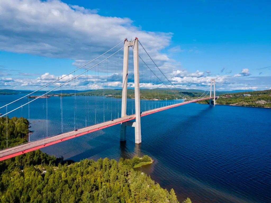 Aerial view of the High Coast Bridge in Sweden spanning a wide blue river surrounded by lush greenery on a sunny day, showcasing Scandinavian engineering and natural beauty.