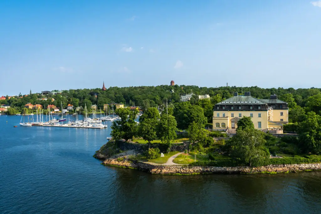 Panoramic view of Djurgarden Island, featuring Skansen Park and yachts docked by the green shoreline of Lake Malaren in Stockholm, Sweden on a sunny day.