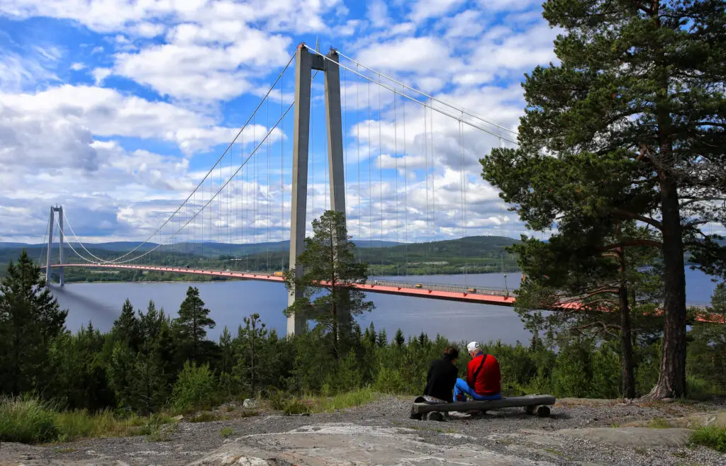 Two tourists enjoying a scenic view of the High Coast Bridge in Sweden, surrounded by lush greenery under a cloudy sky.