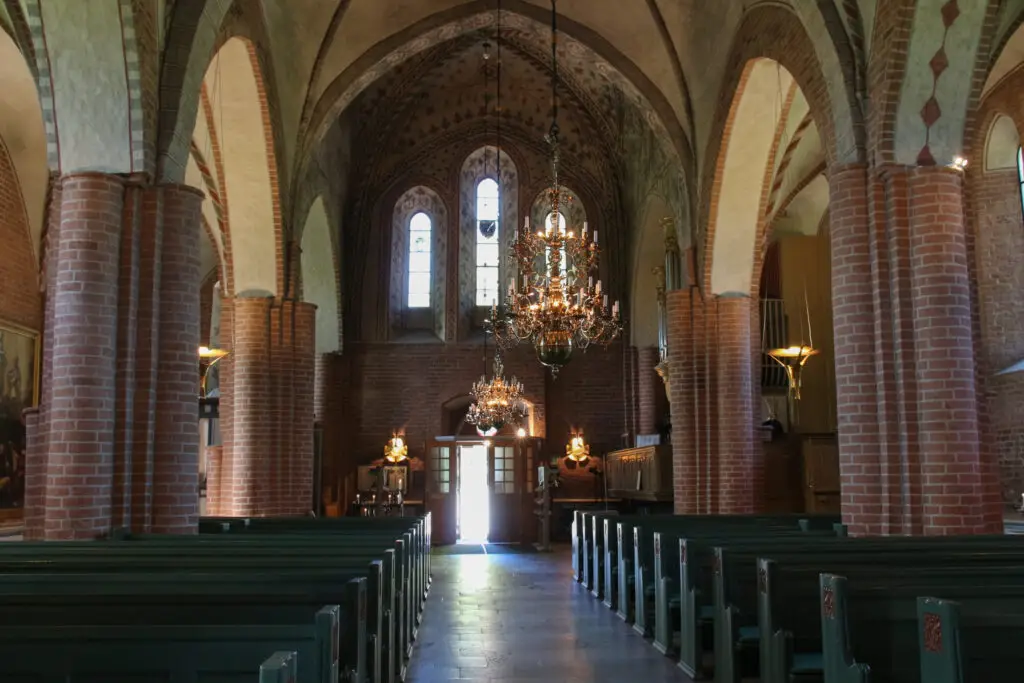 Interior view of Saint Mary's Church (Mariakyrkan) in Sigtuna, Sweden, featuring ornate chandeliers, vaulted ceilings, and brick columns, illuminated by natural sunlight streaming through the entrance.