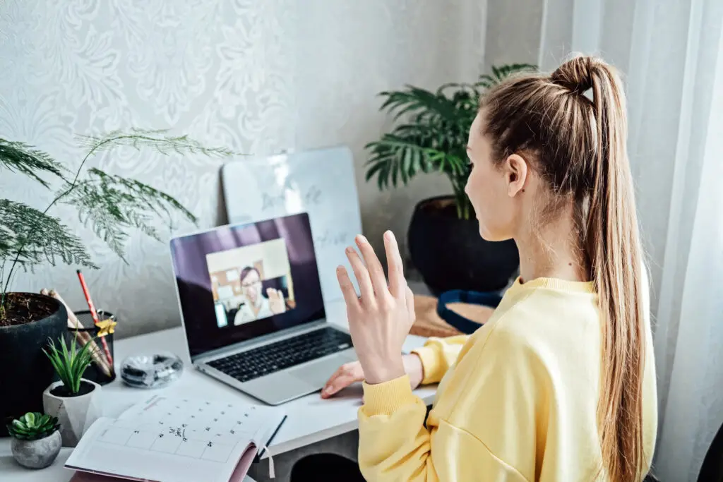 Young woman in yellow sweater engaging in online job interview with female HR manager through video call in a well-organized home office setting.