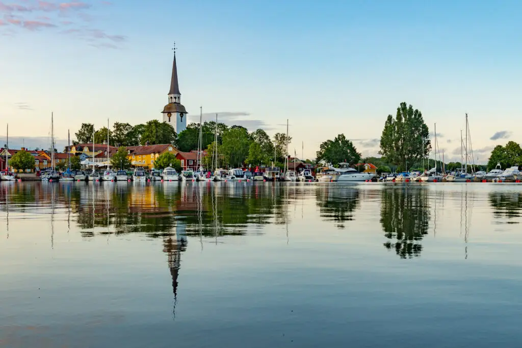 Scenic view of Mariefred, Sweden showing colorful buildings and a church spire reflecting on serene water with boats docked at sunset.