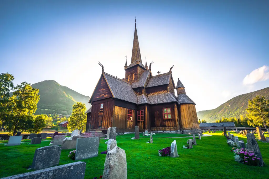 Stave Church of Lom, Norway, an ancient wooden church surrounded by headstones on a sunny day with lush green hills in the background