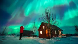 Tourist woman capturing the Northern Lights with her mobile phone in front of a cozy cabin in snowy Iceland landscape.